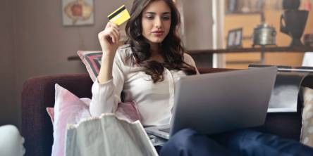 A woman is sitting on the couch looking at something a silver laptop and holding a bank card in her right hand.