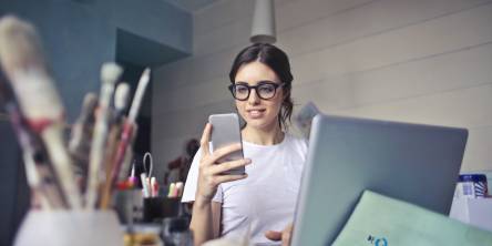 Women sitting behind a craft desk with a laptop. She is looking ah her mobile phone.