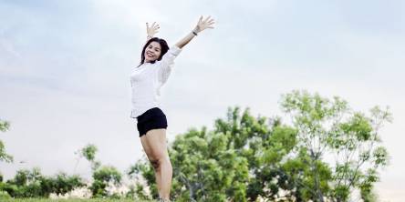 Woman on hill looking happy and content with the sky behind her. Her arms are stretched upward to the sky with a smile.