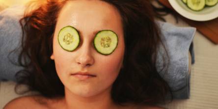 Woman laying down with cucumber slices over her eyes and a bowl of sliced cucumbers by her head on the right hand side.